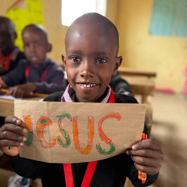 A boy holding up a paper bag he has colored with the name JESUS in orange and green