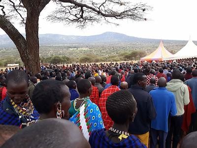 A large crowd of Maasai men and women standing together under acacia trees at Empukani, event tents and hills in the distance