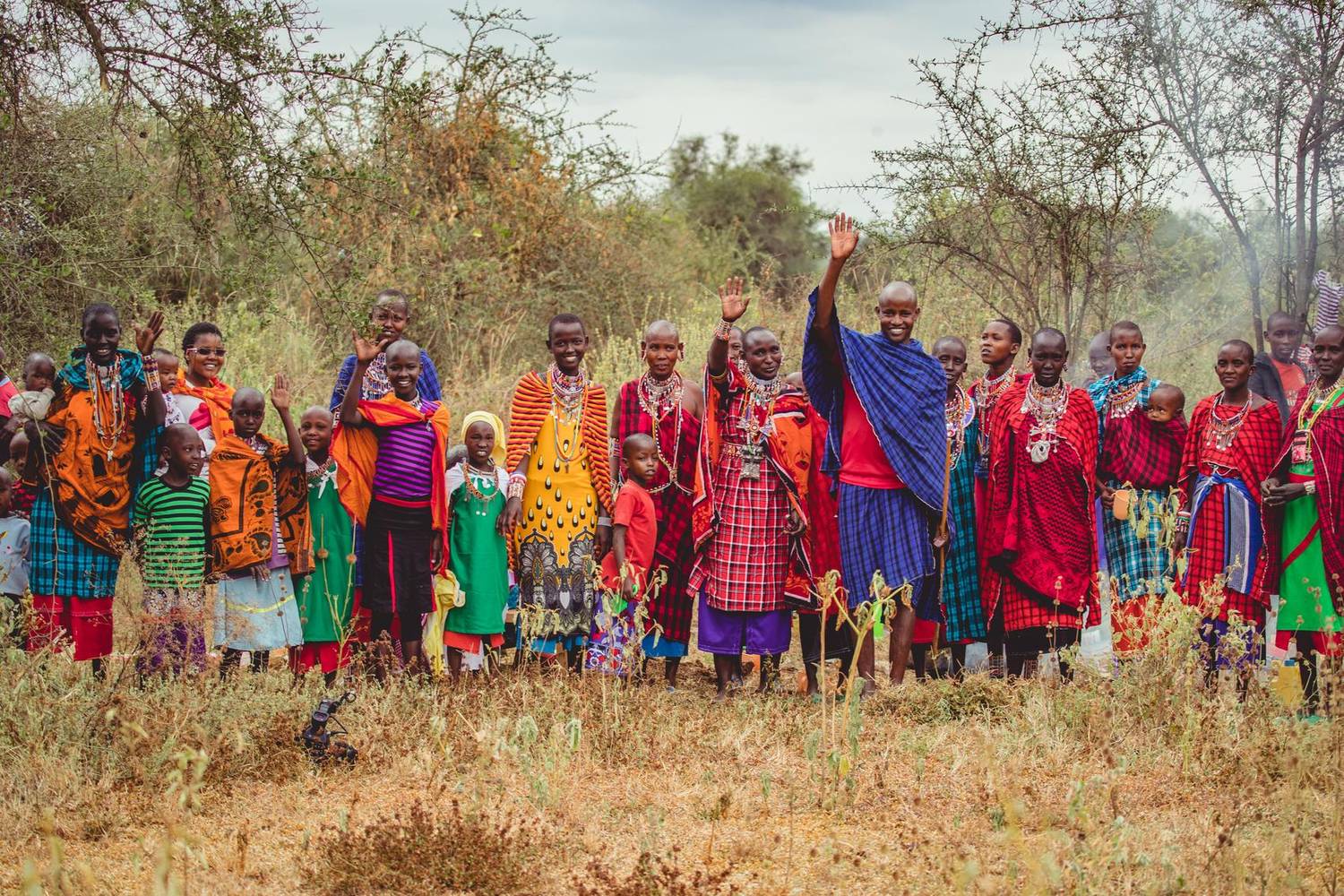 A large group of Maasai women, men and children in bright shukas and beadwork standing together in the bush, several waving and smiling