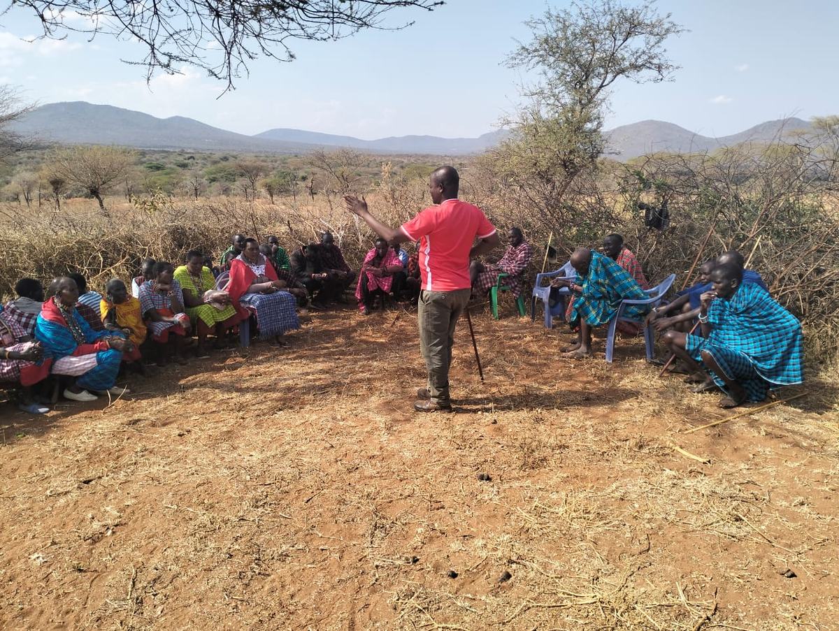 A Maasai evangelist speaking to a seated group of men and women under acacia trees, hills in the distance