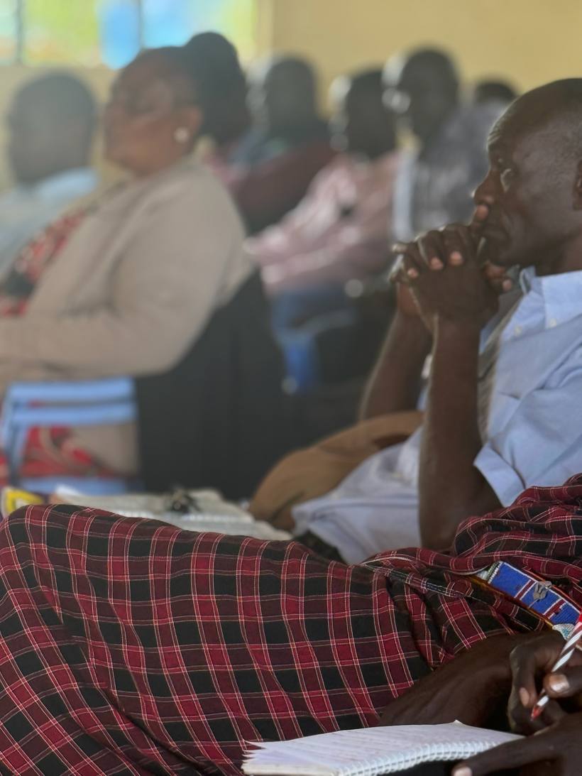 A Maasai pastor in a red shuka listening with his hands clasped, a spiral notebook open on his lap, rows of others seated behind him