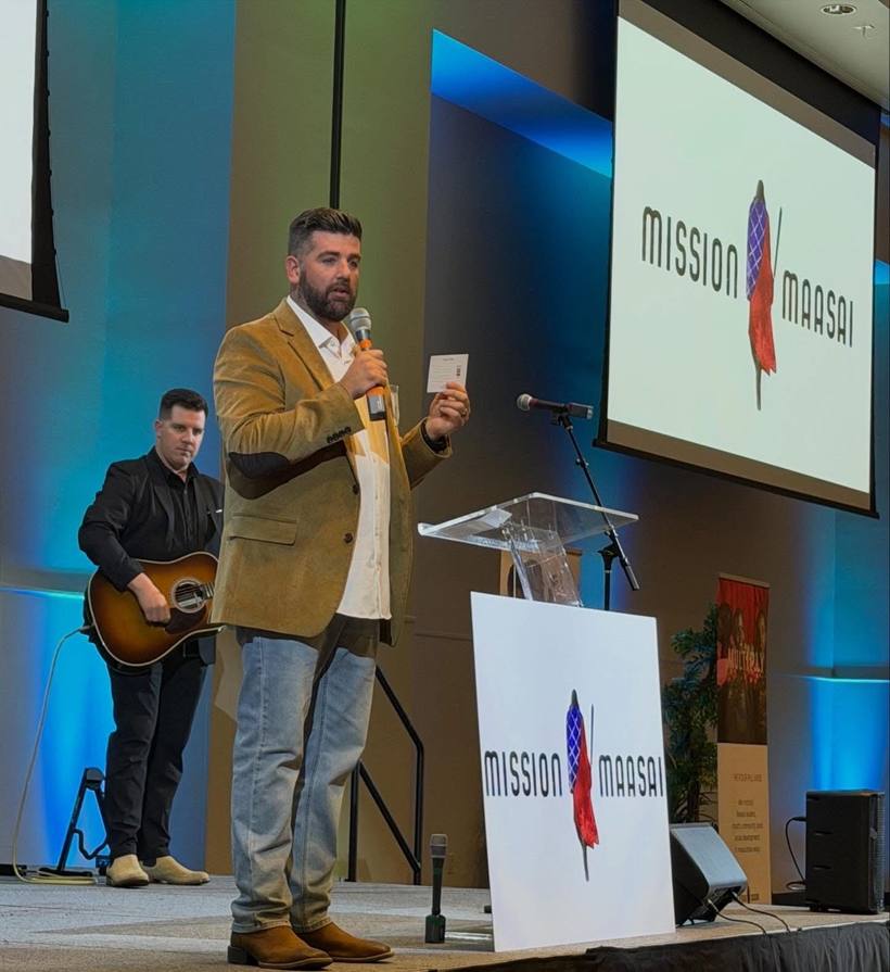 A speaker on stage at the Annual Banquet holding a card, the Mission Maasai logo on the screen behind him and on a board beside the lectern, a guitarist standing to one side