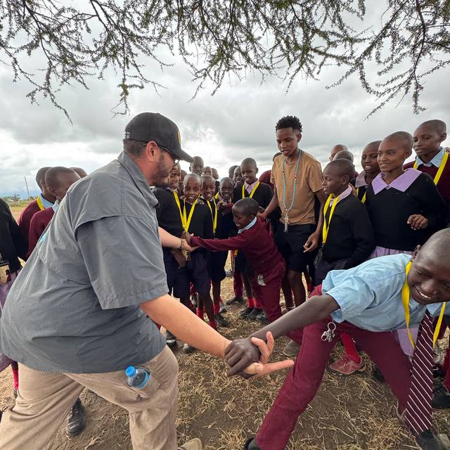 A team member playing a hand-clapping game with students under an acacia tree