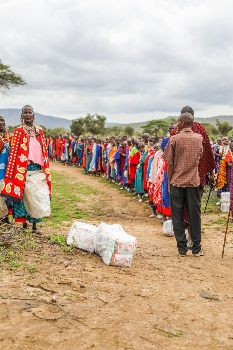 Hundreds of Maasai families queued along a dirt track at a monthly food distribution, sacks of maize flour in the foreground