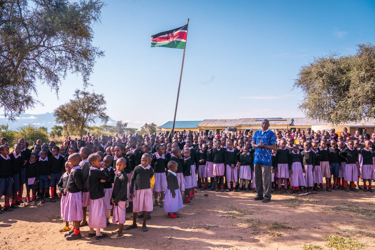 Hundreds of pupils in school uniform lined up for assembly in Empukani beneath the Kenyan flag, a teacher standing among them