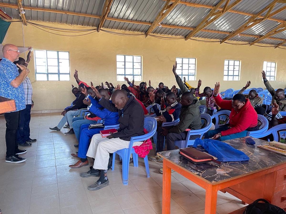 Seminary students seated in a classroom with hands raised in prayer, Bibles on their laps, a teacher standing at the front