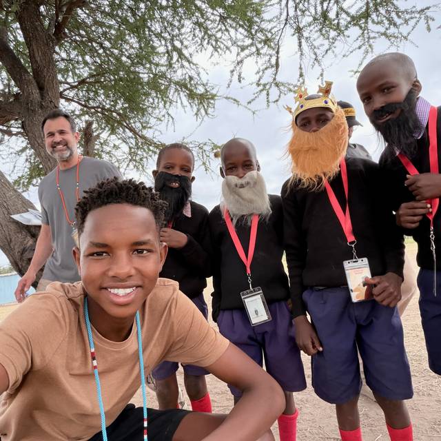 Students wearing costume beards for a Bible story, laughing under an acacia tree