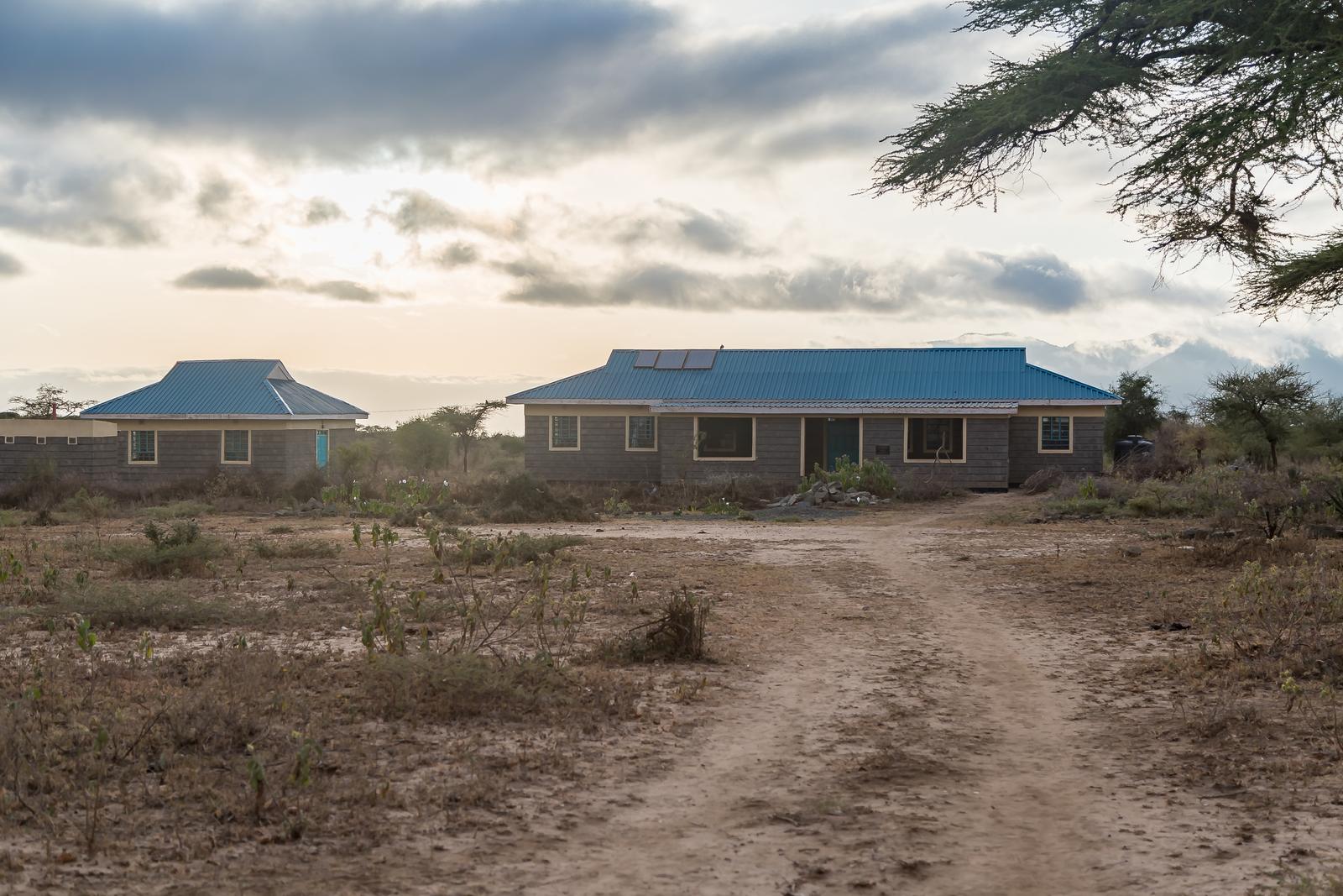 The Empukani Medical Clinic at dusk, two block buildings with blue metal roofs and solar panels, a dirt track leading up to the door across dry ground