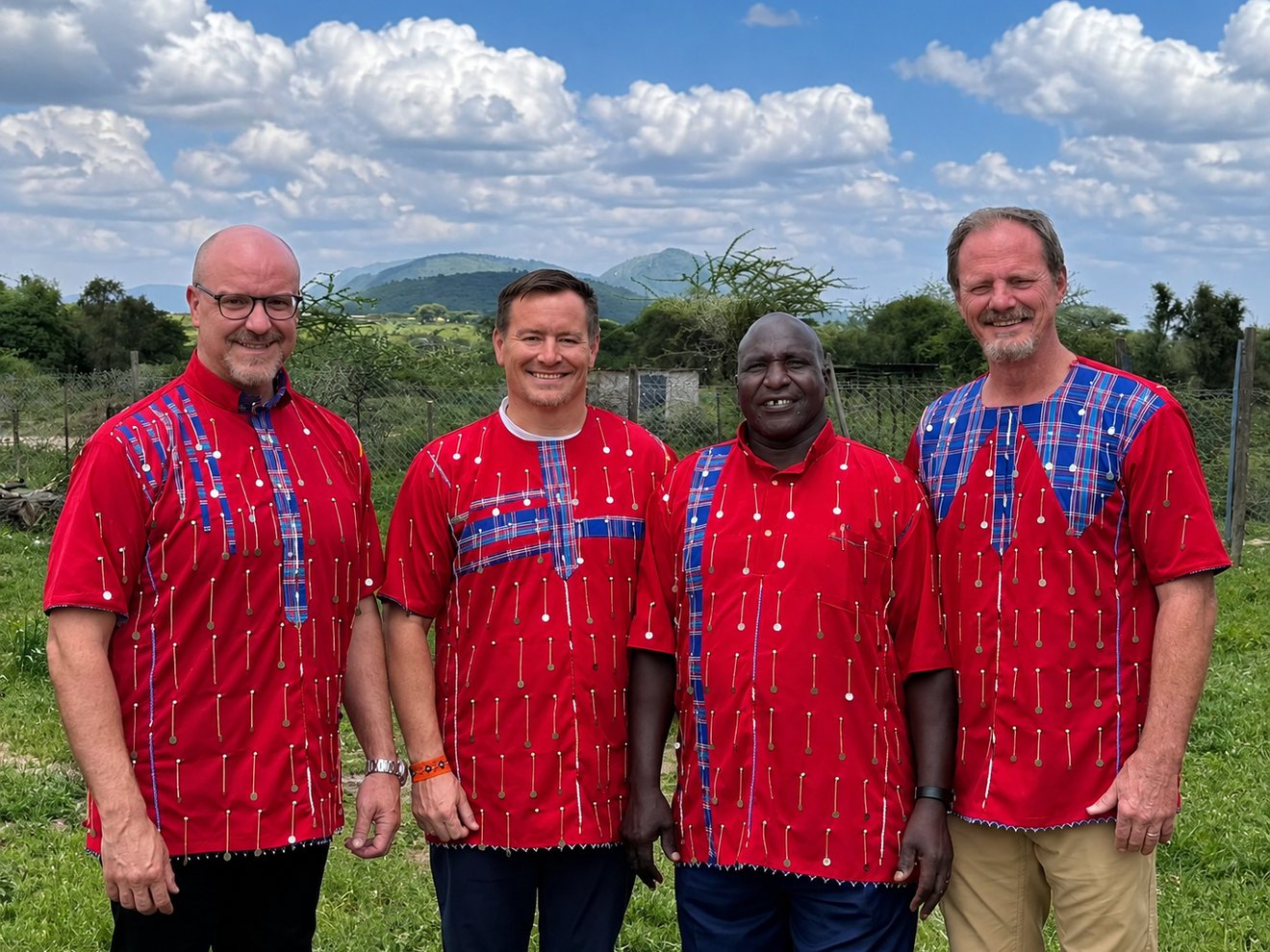 The four members of the Mission Maasai board standing together on grass in red Maasai shirts, a fence line and green hills behind them
