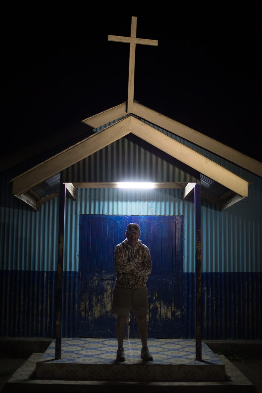 The original Empukani church at night, corrugated iron walls painted blue, a wooden cross on the roofline, a single light over the doorway