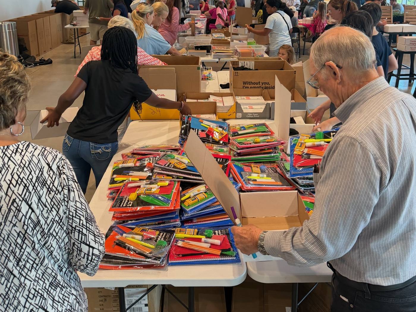 Volunteers of all ages around long tables, packing stacks of notebooks, pencils and supplies into boxes for the children in Empukani